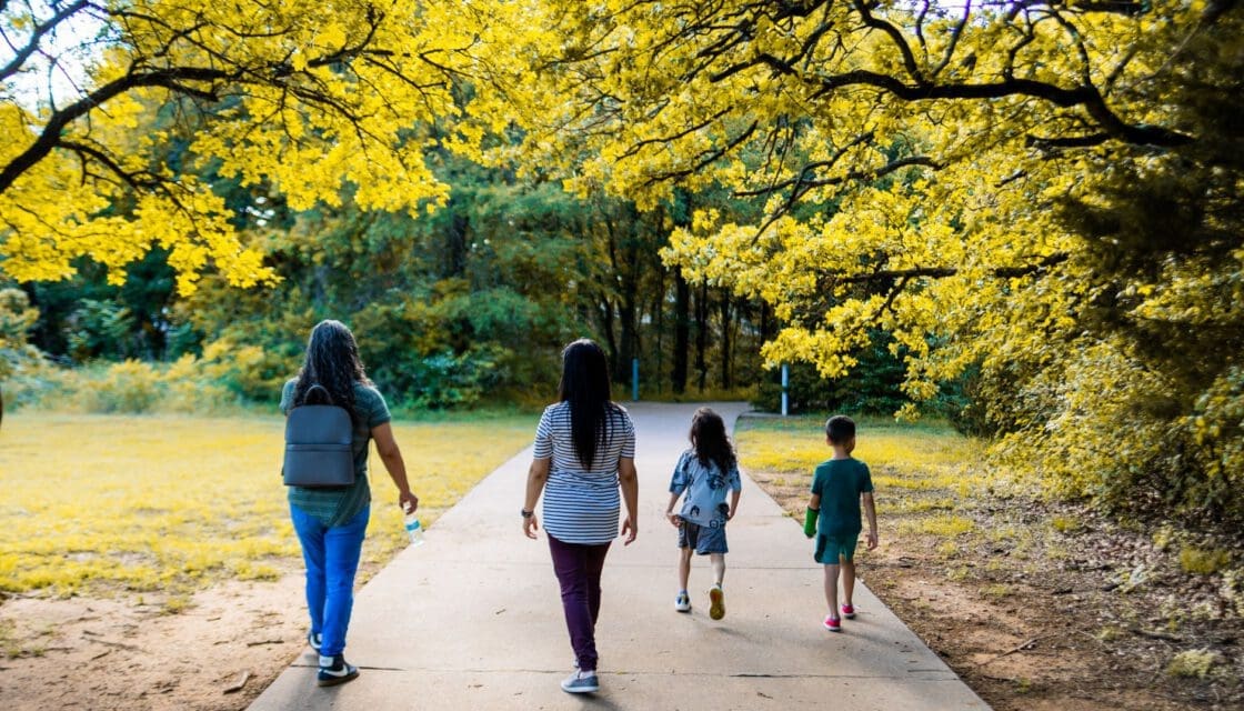 Brackenridge Park San Antonio stroller — women and children walking along a tree-lined park pathway in golden light